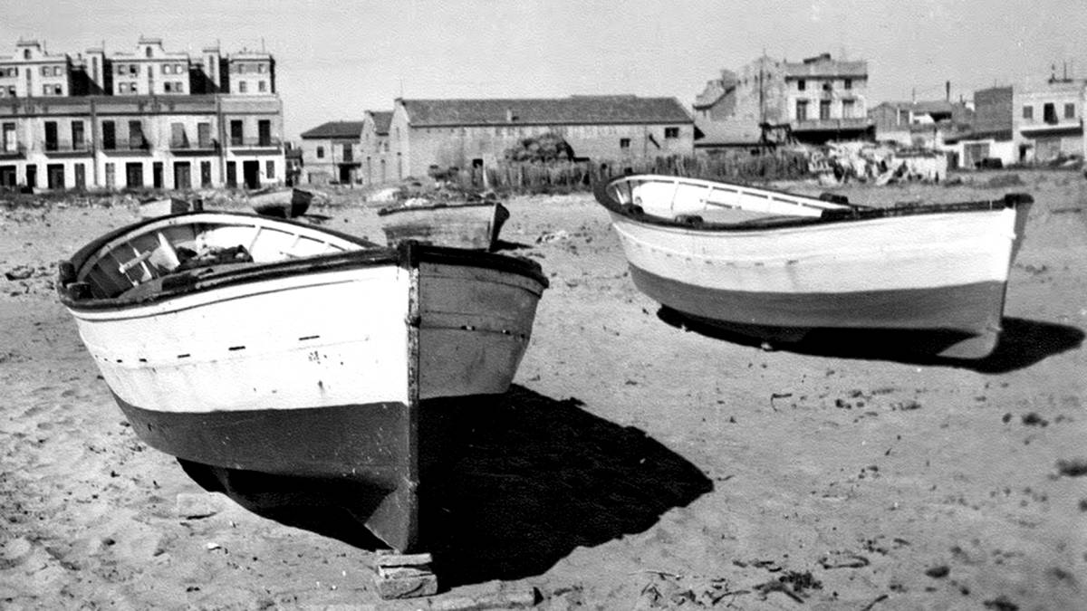 Barques varades en la platja, davant les dunes. Darrere, la Fàbrica de Gel i els blocs de portuaris